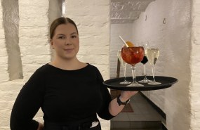 Tarjoilijaa pitelee tarjotinta, jossa on täysiä valkoviinilaseja sekä yksi iso drinkki. Taustalla Kellariravintola Hällän tiiliseinää. 