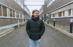 Jimi Leino seisomassa HAMKin kampuksella Riihimäellä ulkona kahden tiilirakennuksen seinien välissä kulkevalla hiekkakäytävällä