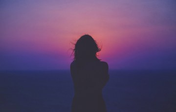 Naisen puolikas siluetti punertavanviolettia auringonlaskua vasten. Taustalla utuinen horisontti.