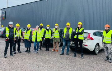 Opiskelijoita keltaisissa turvaliiiveissä ja kypärät päässä Versowoodin tehtaan edustalla ulkona.
