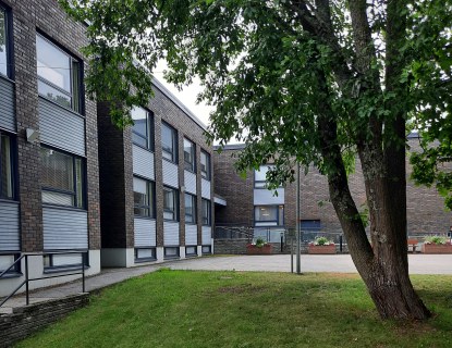 Kaksikerroksisen, L:n muotoisen tiiliverhoillun Kauniston palvelukeskuksen sisäpiha kesällä, oikealla etualalla iso lehtipuu, taustalla talon edustalla istutuksia