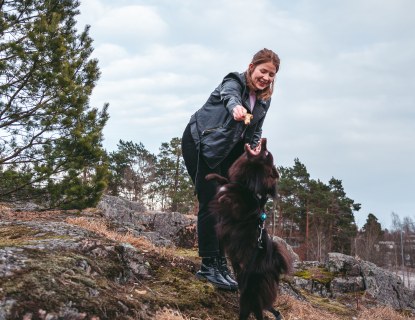 Nainen ja koira leikkimässä kalliolla. Taustalla mäntyjä ja taivasta. 