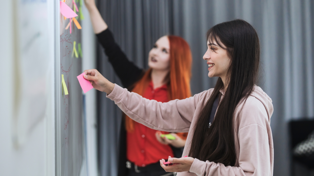 Valokuva kahdesta henkilöstä, jotka kiinnittävät post-it -lappuja valkotaululle. Henkilöt hymyilevät.